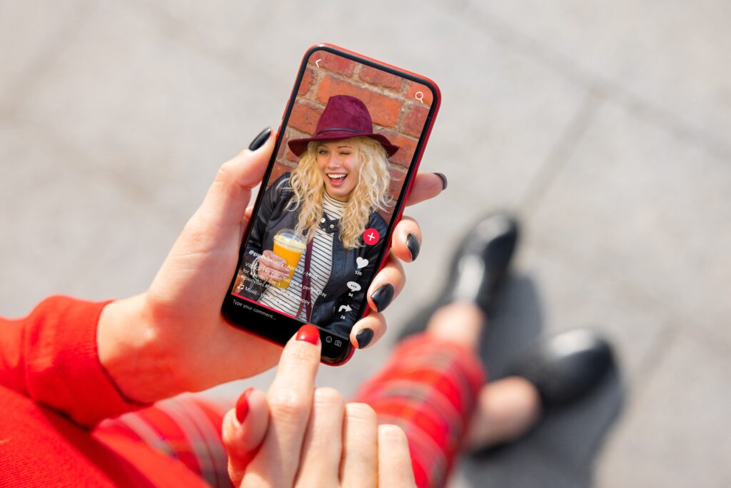 Pessoa segurando um smartphone exibindo um vídeo do TikTok com uma mulher sorridente de chapéu roxo, segurando uma bebida. A usuária interage com o vídeo, que mostra curtidas, comentários e botões de interação típicos da plataforma. Cena representa o consumo de conteúdo digital e a influência das redes sociais na comunicação moderna.