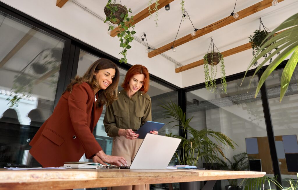 Duas mulheres profissionais analisando campanhas de marketing em um ambiente moderno com vegetação, usando notebook e tablet em mesa de trabalho.