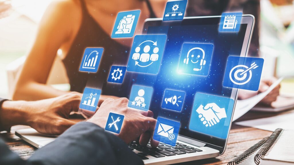 Profissionais trabalhando em um notebook com ícones digitais flutuando na tela, representando estratégias de marketing digital como e-mail, CRM, funil de vendas, automação, atendimento, análise de dados e metas. A cena simboliza o uso da tecnologia e dados para alavancar resultados empresariais, com foco em tendências de marketing digital 2025.